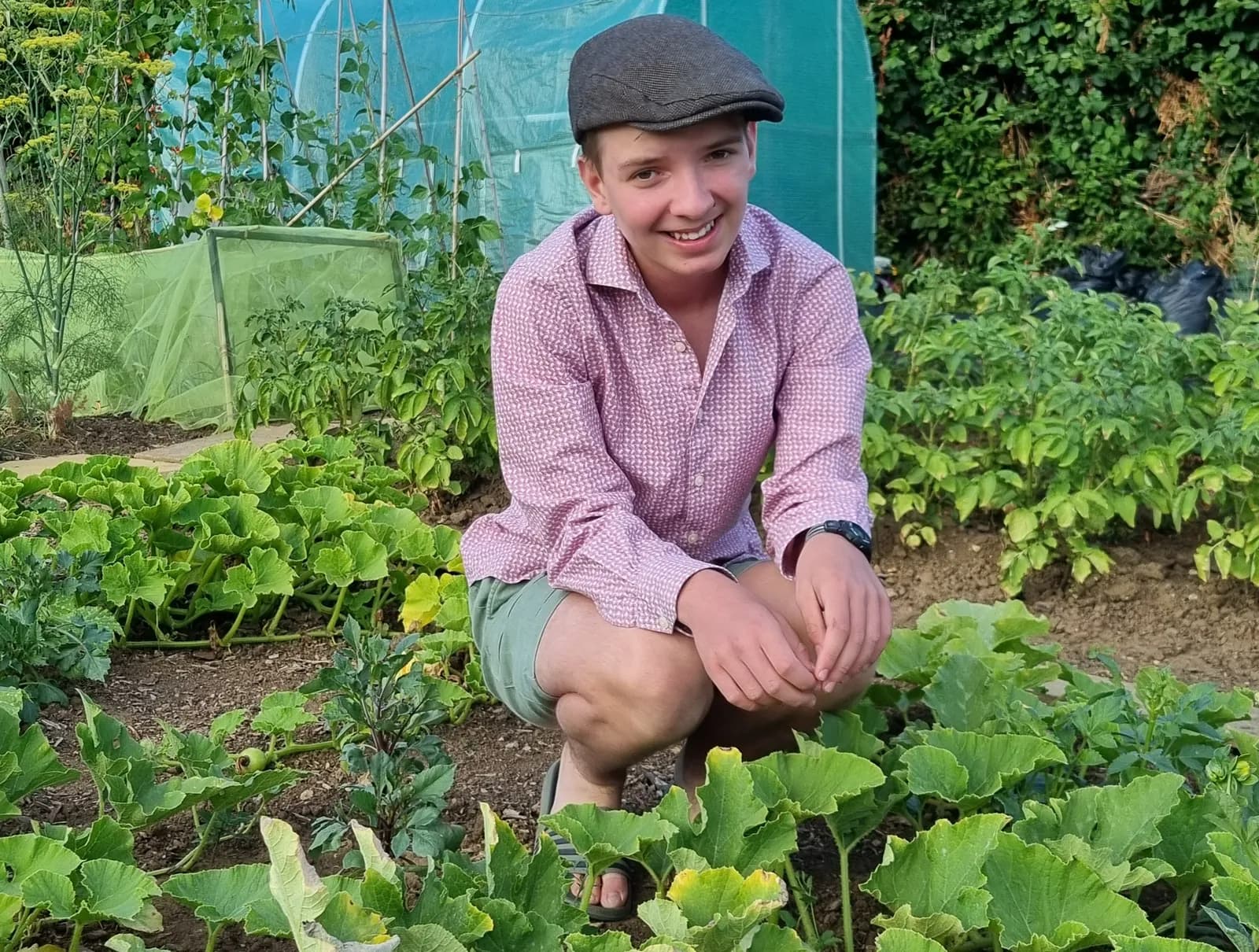 A young person wearing a flat cap and a pink patterned shirt smiles while crouching in a garden among green plants and a greenhouse in the background.